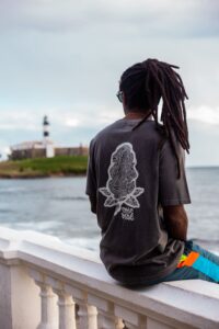 Camisa cor chumbo washed com marca da CrocBus em branco em modelo sentado frente ao mar com o Farol da Barra ao fundo